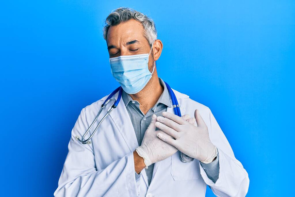 Hombre de pelo gris de mediana edad vistiendo uniforme médico y máscara médica sonriendo con las manos en el pecho, los ojos cerrados con gesto agradecido en la cara. concepto de salud.  - Foto, Imagen