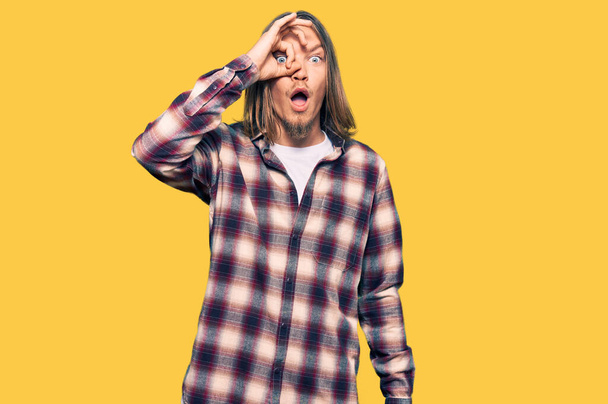 Handsome caucasian man with long hair wearing hipster shirt doing ok gesture shocked with surprised face, eye looking through fingers. unbelieving expression.  - Fotografie, Obrázek