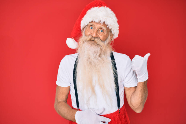 Старик с седыми волосами и длинной бородой в белой футболке и костюме Санта-Клауса улыбается со счастливым лицом, смотрящим и указывающим на сторону с большим пальцем.  - Фото, изображение