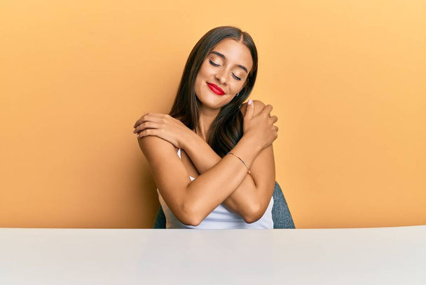 Fiatal spanyol nő alkalmi ruhát visel ül az asztalon öleli magát boldog és pozitív, mosolygós magabiztos. önszeretet és öngondoskodás  - Fotó, kép