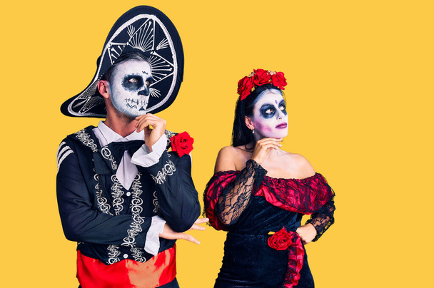 Молодая пара, одетая в мексиканский день мертвого костюма на заднем плане с рукой на подбородке, думает о вопросе, задумчивом выражении лица. улыбаясь с вдумчивым лицом. Концепция сомнений.  - Фото, изображение