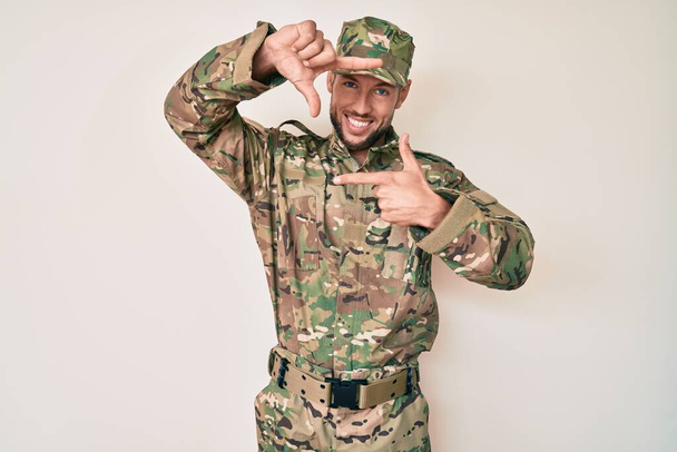 Young caucasian man wearing camouflage army uniform smiling making frame with hands and fingers with happy face. creativity and photography concept.  - Фото, зображення