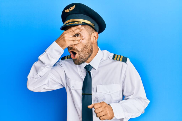 Handsome man with beard wearing airplane pilot uniform peeking in shock covering face and eyes with hand, looking through fingers with embarrassed expression.  - Fotografie, Obrázek
