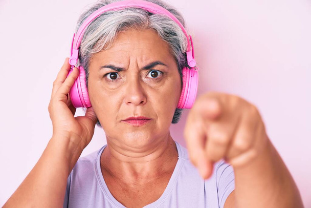 Starsza Latynoska słuchająca muzyki przy użyciu słuchawek wskazujących palcem na kamerę i na ciebie, pewny siebie gest wyglądający poważnie  - Zdjęcie, obraz