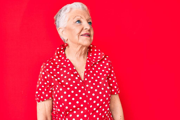 Senior schöne Frau mit blauen Augen und grauen Haaren trägt lässige Sommerkleidung über rotem Hintergrund zur Seite schauen, entspannen Profil Pose mit natürlichem Gesicht und selbstbewusstem Lächeln.  - Foto, Bild