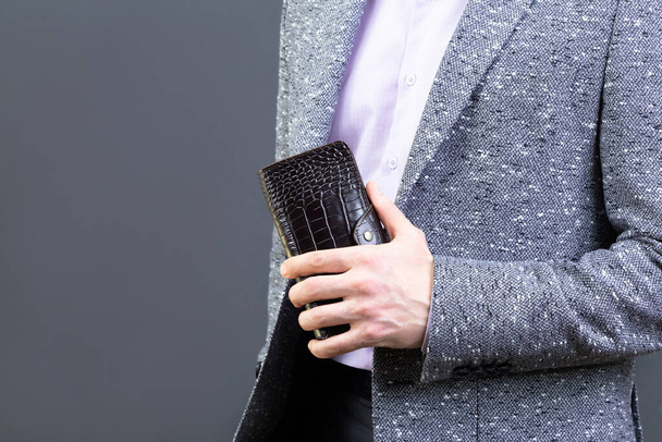 Homem de casaco cinzento segurando uma carteira de couro marrom, close-up. Fundo cinzento neutro. Conceito de moda e compras - Foto, Imagem