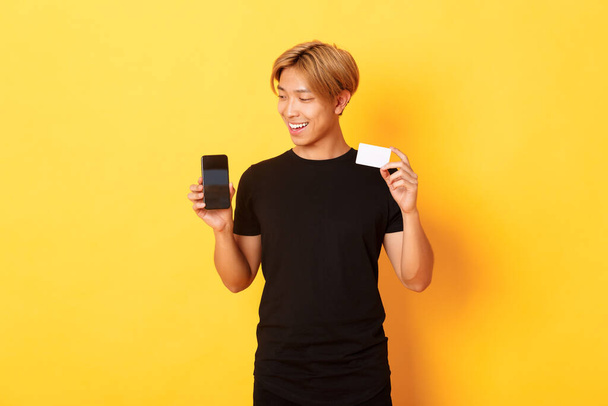 Retrato de chico asiático guapo complacido, mostrando la pantalla del teléfono inteligente y la tarjeta de crédito, mirando la pantalla del teléfono móvil, fondo amarillo - Foto, Imagen