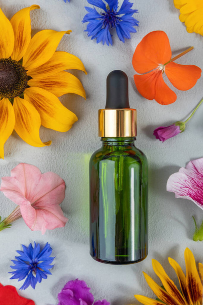 Anti-aging collagen, facial serum or other cosmetic product in green glass bottle among the colored flowers on grey background. Natural Organic Spa Cosmetic concept Mockup Top view. - Foto, imagen
