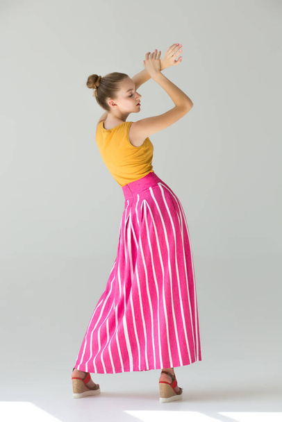 The girl stands with her back in full growth, her arms are raised above, her eyes are closed. Clothes - long red skirt in wide white striped and orange tank top. Summer look. - Foto, Imagen