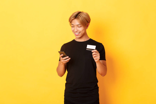 Sonriente guapo asiático hombre pagar por la compra en línea, compras en Internet, la celebración de teléfono inteligente y tarjeta de crédito, fondo amarillo - Foto, Imagen
