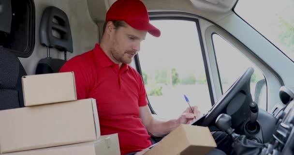 jakelupalvelun mies istuu pakettiautossa ja kirjoittaa asiakirjoja leikepöydälle ennen paketin toimittamista asiakkaalle - Materiaali, video