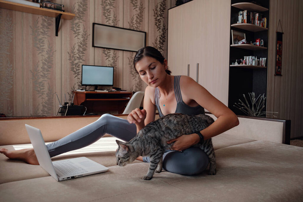 Young attractive smiling woman practicing yoga, sitting in Half Lotus exercise, wearing sportswear, meditation session, indoor full length, home interior, cat near. - Photo, Image