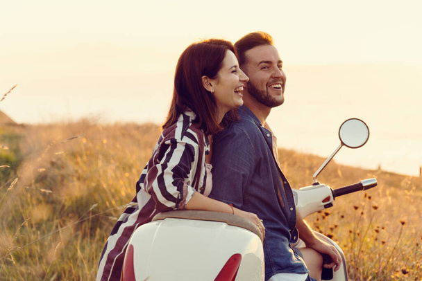Пара молодых друзей в свежей одежде, веселятся и улыбаются на мотоцикле на побережье во время заката солнца - Фото, изображение
