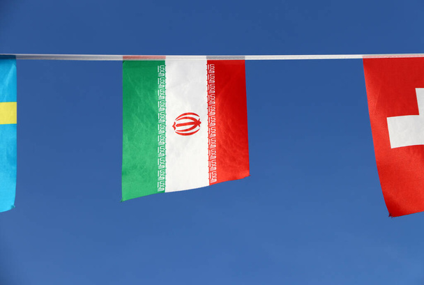 Mini-Schienenfahne des Iran in Trikolore grün-weiß-rot mit Nationalwappen in roter Mitte auf dem weißen Band und dem Takbir, der 11 Mal in der Kufic-Schrift in weiß geschrieben wurde, auf jedem Band, das auf dem Seiltuch zwischen der Flagge anderer Länder hängt - Foto, Bild