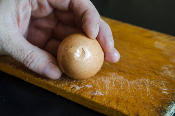 Una mano masculina sostiene un huevo de gallina hervido en su cáscara contra la tabla de cortar. Método de limpieza de las conchas. Cocinar. Enfoque selectivo. - Foto, Imagen