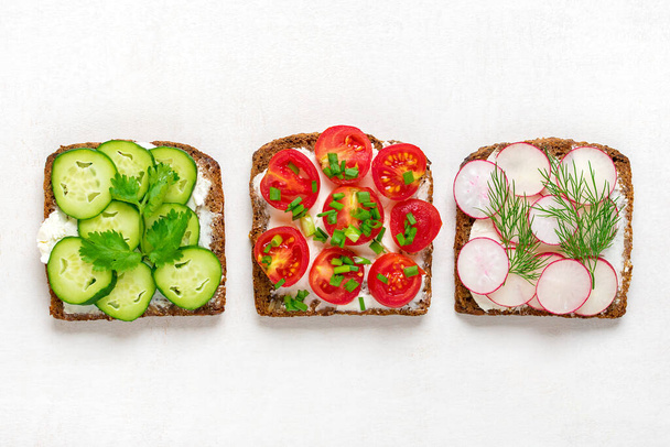 Variety of sandwiches for breakfast with slice of whole grain dark bread, cream cheese, cucumbers, radishes, cherry tomatoes, garnished with dill, green onions on light white table Top view Flat lay. - Photo, Image