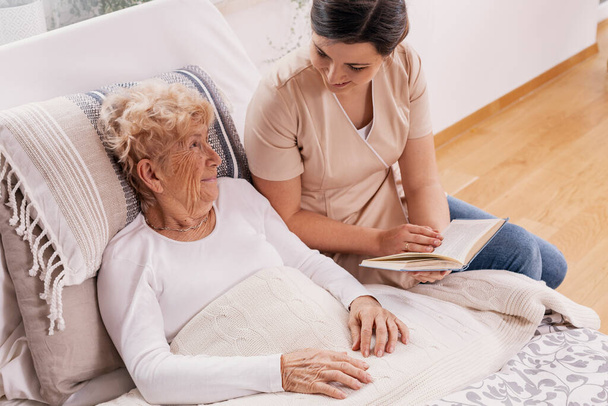 Helpful female volunteer reads a book to an elderly woman lying in a bed - Foto, Bild