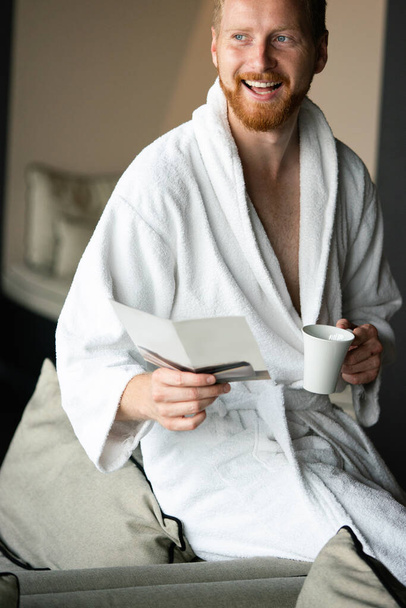 Jovem homem bonito feliz bebendo café da manhã em roupão de banho - Foto, Imagem
