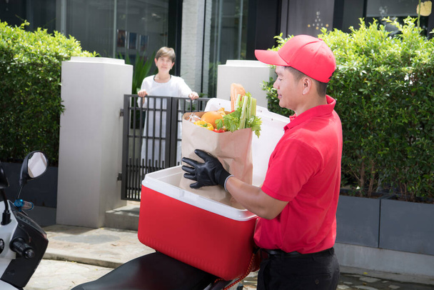 Kırmızı üniformalı Asyalı teslimatçı eve yiyecek, meyve, sebze ve içecek getiren kadına yiyecek getiriyor. - Fotoğraf, Görsel
