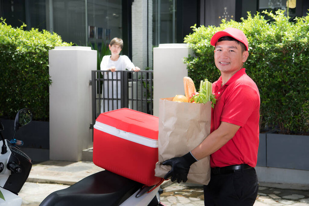 Asiatischer Zusteller in roter Uniform liefert Lebensmittel, Obst, Gemüse und Getränke an Empfängerin zu Hause - Foto, Bild