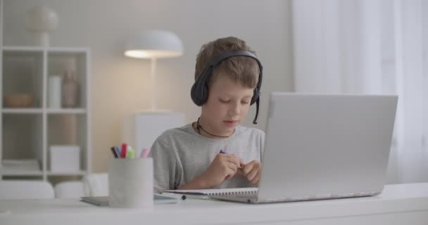 Маленький мальчик рисует войлочные ручки, сидя дома в выходные, слушая аудио в наушниках, ноутбук перед ним - Кадры, видео