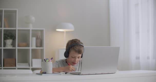 σχολείο αγόρι ακούει παραμύθι μέσω ακουστικών από σημειωματάριο και σχέδιο, κάθεται στο τραπέζι μόνη της στο δωμάτιο - Πλάνα, βίντεο