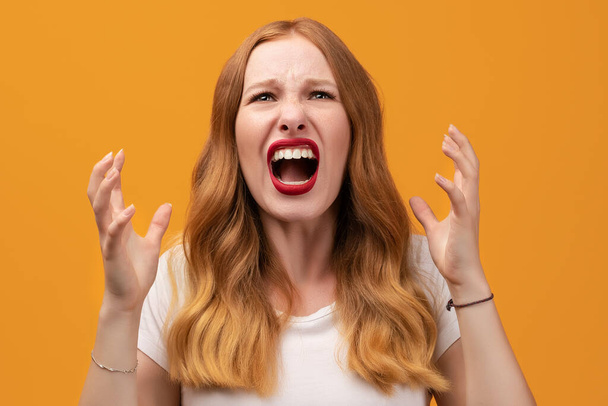 Angry young woman with wavy redhead, wearing white t-shirt screaming on yellow studio background. Emotional face. Human emotions, facial expression concept. - Photo, Image