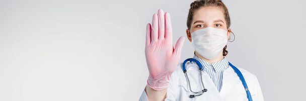 Una joven y hermosa doctora con una bata blanca y guantes estériles rosados muestra un gesto de stop sobre fondo blanco. No digas enfermedad por virus. Copia espacio para tu texto - Foto, imagen