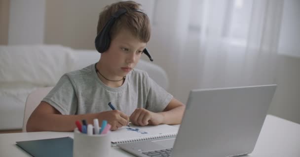 kotiopetuksessa klo pandemia koronaviruksen, pieni poika kuuntelee opettaja kuulokkeilla ja kannettavan tietokoneen, piirustus paperille - Materiaali, video