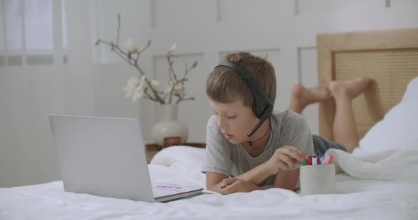 Мальчик-подросток раскрашивает и рисует картины в альбоме, лежа дома на кровати и общаясь с помощью ноутбука - Кадры, видео