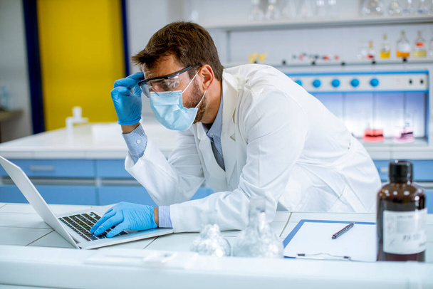 Όμορφος νέος ερευνητής σε λευκό εργαστηριακό παλτό και προστατευτική μάσκα εργασίας χρησιμοποιώντας φορητό υπολογιστή, ενώ κάθεται στο εργαστήριο - Φωτογραφία, εικόνα