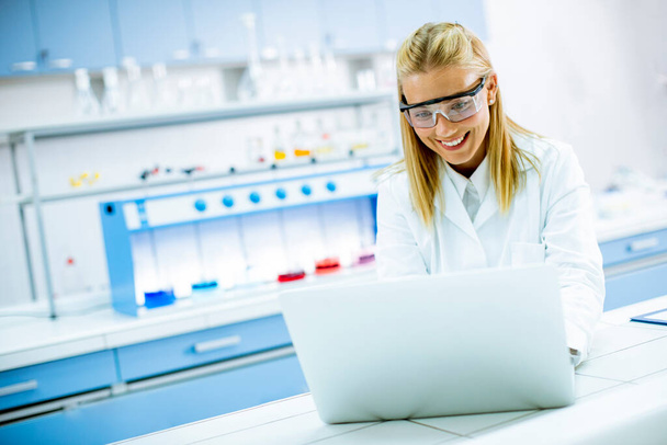 Χαριτωμένη ερευνήτρια με λευκή ρόμπα εργαστηρίου και προστατευτικά γυαλιά που χρησιμοποιούν φορητό υπολογιστή ενώ εργάζονται στο εργαστήριο - Φωτογραφία, εικόνα