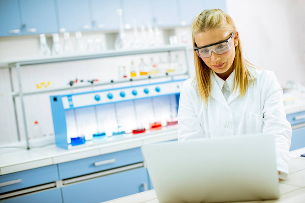 Linda investigadora en bata de laboratorio blanca y gafas protectoras usando portátil mientras trabaja en el laboratorio - Foto, imagen