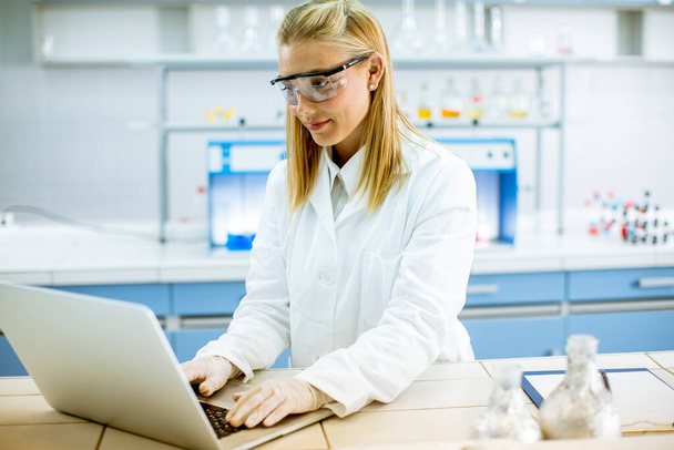 Симпатичная женщина-исследователь в белом халате и защитных очках с ноутбуком во время работы в лаборатории - Фото, изображение
