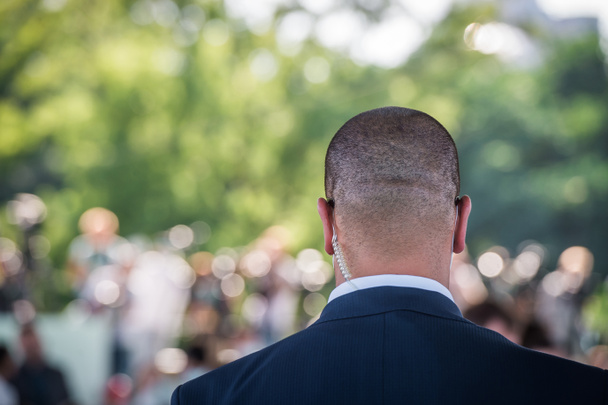 Security guard listening to his earpiece on event. Back of jacket showing. secret service guard. private bodyguard. man with earpiece in crowd. Black suit and bald head. - Photo, Image