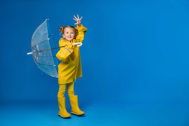 Cheerful girl in a raincoat and rubber boots standing on a blue background with a transparent umbrella. Copy space picture with placement for your text of advertisement - Photo, Image
