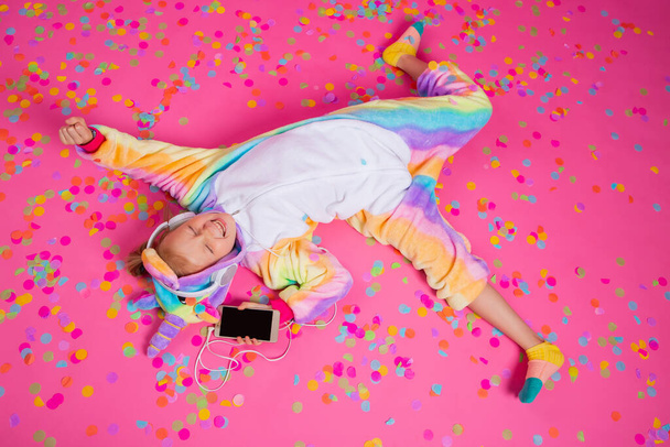 Happy little blonde girl in unicorn unitard listens to music from her smartphone.  Isolated on pink background with a space for text - Photo, Image