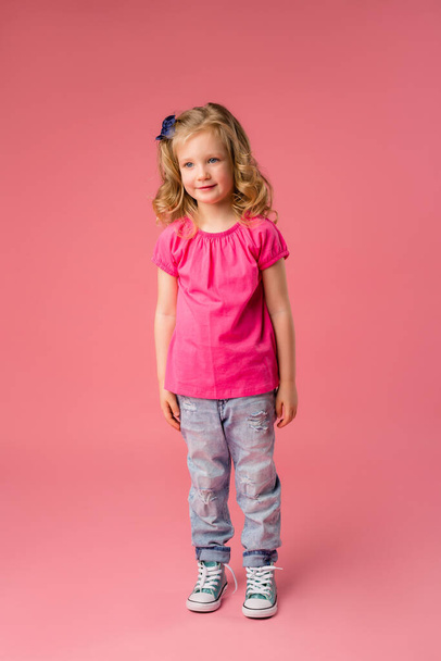 Roztomilá malá blondýnka v růžovém tričku a džínách na růžovém pozadí. Kopírovat mezerník s umístěním pro váš text nebo inzerát - Fotografie, Obrázek