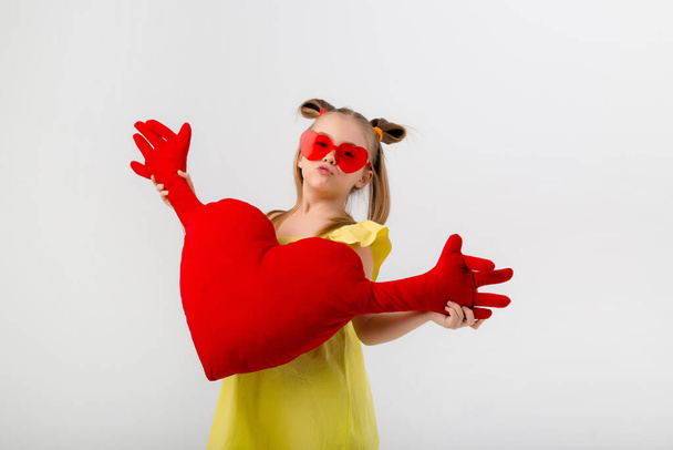 Happy girl in heart shaped sunglasses hugs a red heart pillow. Isolated on white background with a space for text  - Photo, Image
