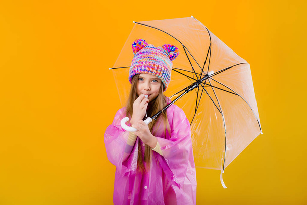 Photo of a little girl in a pink raincoat and  knitted hat holding an umbrella isolated on yellow background with a space for text - Фото, изображение