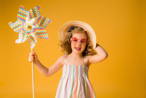 Μικρό χαρούμενο κορίτσι με μεγάλα γυαλιά ηλίου και ψάθινο καπέλο που κρατάει μια ρόδα σε κίτρινο φόντο. Αντιγραφή εικόνας χώρου με τοποθέτηση για το κείμενο ή τη διαφήμισή σας - Φωτογραφία, εικόνα
