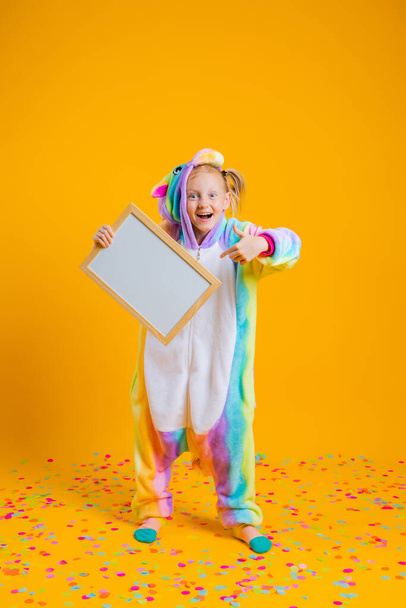 Una bambina felice in una tuta kigurumi unicorno tiene una tavola vuota per il testo su uno sfondo giallo. Concetto vacanza o informazioni sugli sconti - Foto, immagini
