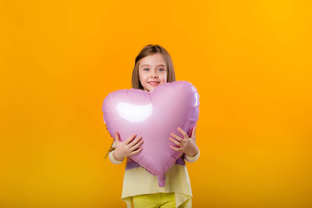 Retrato de uma criança feliz menina sorrindo e segurando um balão rosa na forma de um coração. Isolado em fundo amarelo com um espaço para texto - Foto, Imagem