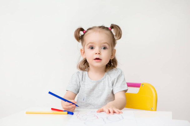 Foto eines zweijährigen Mädchens sitzt an einem Tisch und zeichnet mit bunten Filzstiften auf weißem Hintergrund - Foto, Bild