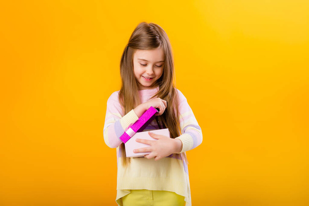 Foto van een gelukkig klein meisje met een roze geschenkdoos, geïsoleerd op gele achtergrond met een ruimte voor tekst - Foto, afbeelding