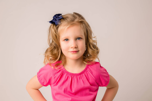 Portrait d'une petite fille blonde aux boucles dans un t-shirt rose sur fond beige - Photo, image
