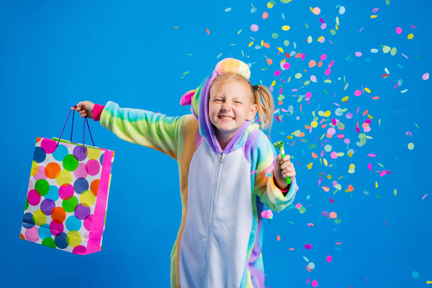 Tek boynuzlu at kostümü giymiş mutlu küçük bir kız elinde renkli konfeti dolu bir hediye çantası tutuyor. Metin için boş alanı olan doğum günü, tatil veya satış kavramı - Fotoğraf, Görsel