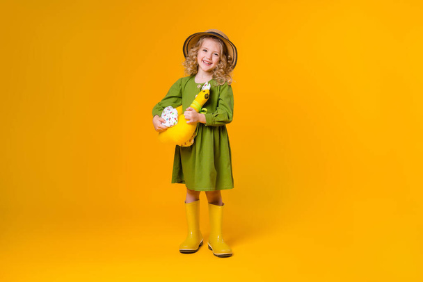 Little girl in a green linen dress and yellow rubber boots holding a textile toy chicken on a yellow background. Copy space picture with placement for your text or advertisement, concept of eco-friendly lifestyle - Фото, изображение