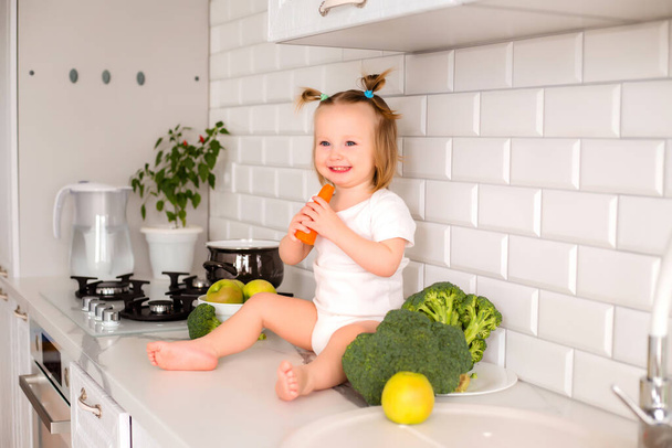 Foto de una niña sentada en la mesa de la cocina. Un niño juega y se divierte con verduras y frutas orgánicas frescas en la cocina blanca crujiente - Foto, Imagen