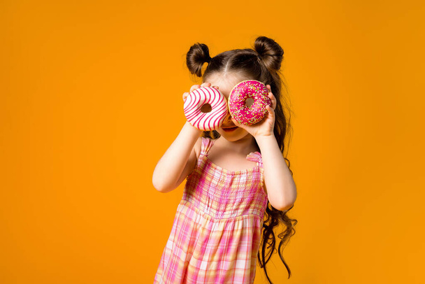 Cheerful girl having fun with doughnuts. Isolated on orange background, children's emotions concept. - Fotografie, Obrázek